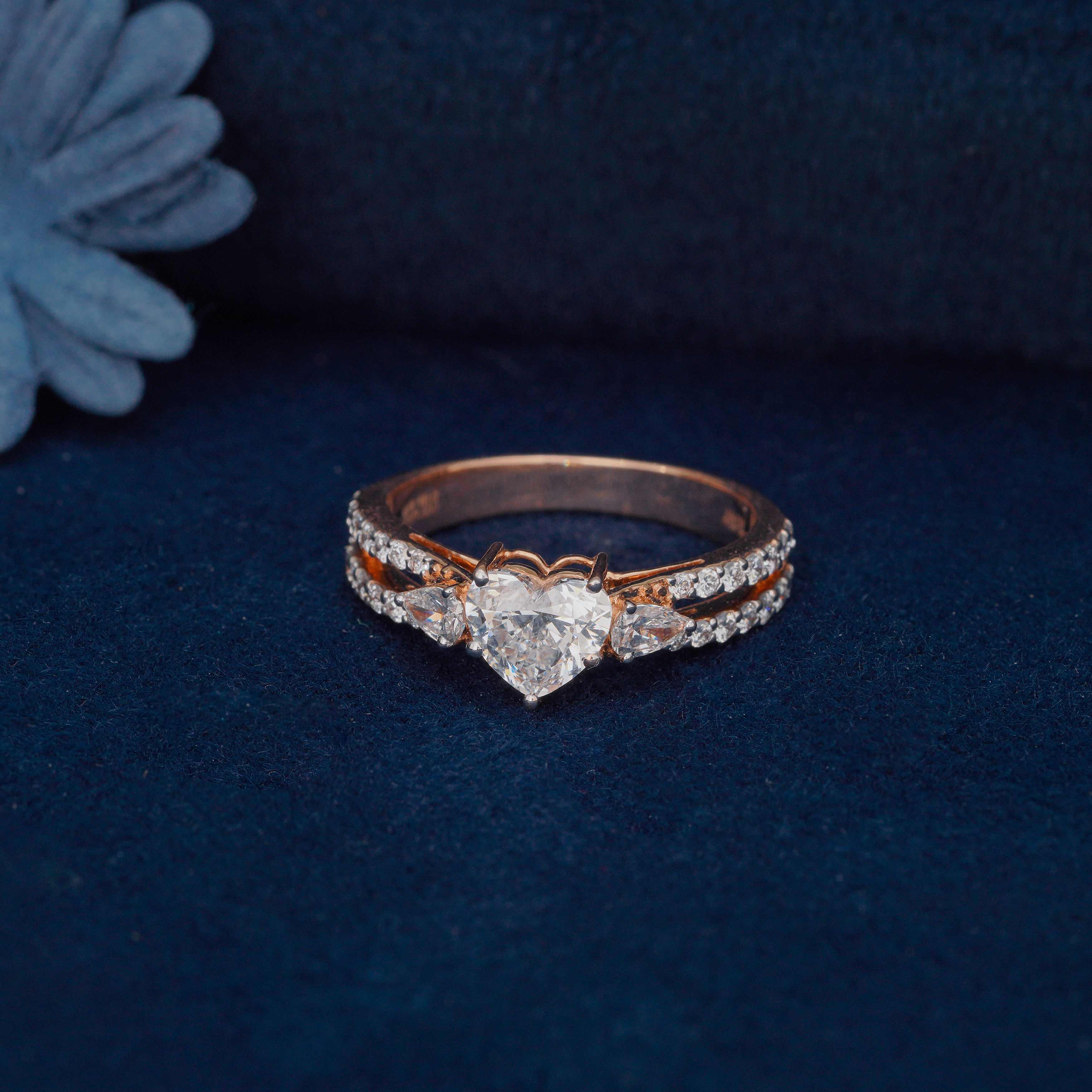 Heart-shaped diamond ring with side stones on a double-row pavé yellow gold band, set against a dark blue background.