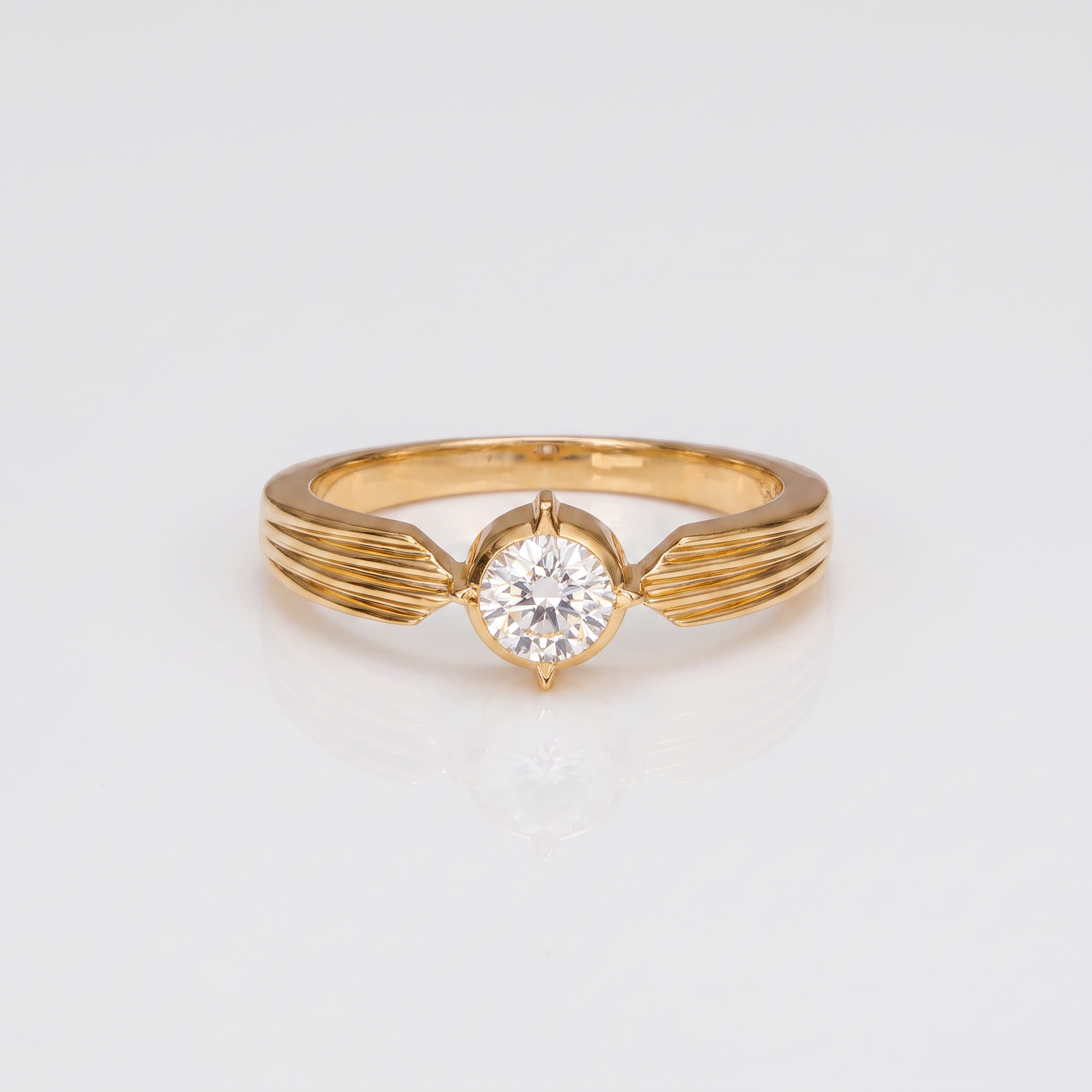 A yellow gold solitaire ring with a round-cut clear gemstone, featuring a wing-like twisted band design, shown on a clean white background with a soft reflection.
