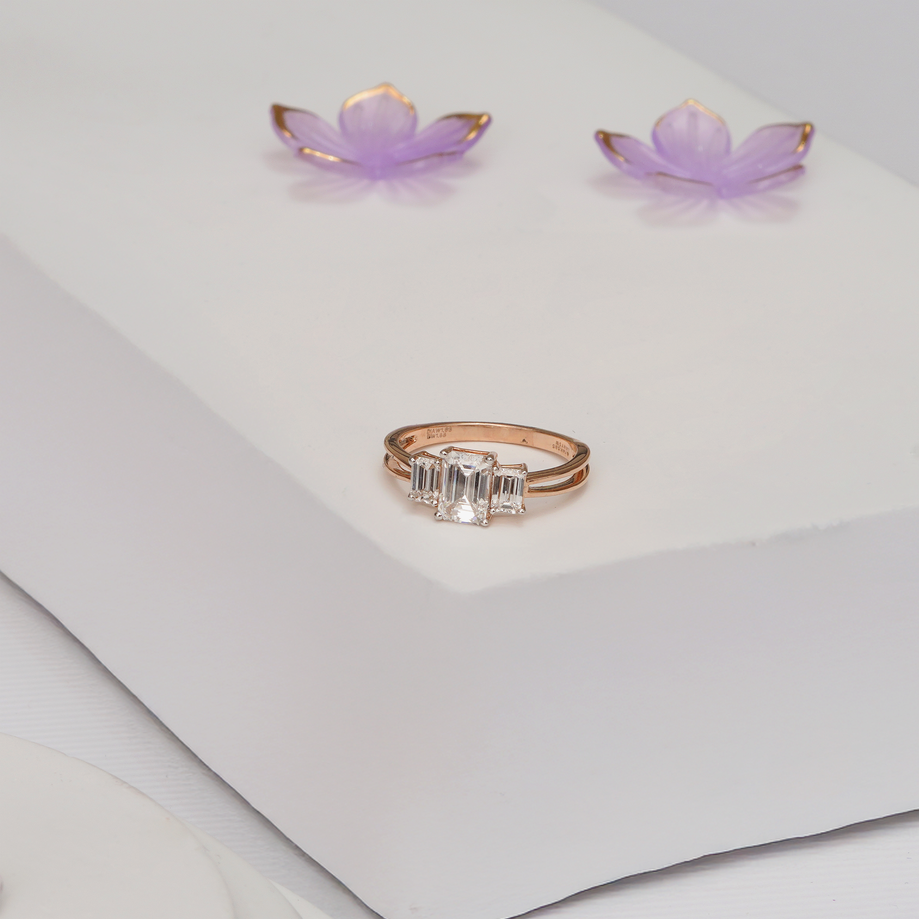Three-stone emerald-cut diamond ring in 18k yellow gold on a white display stand with purple flowers.
