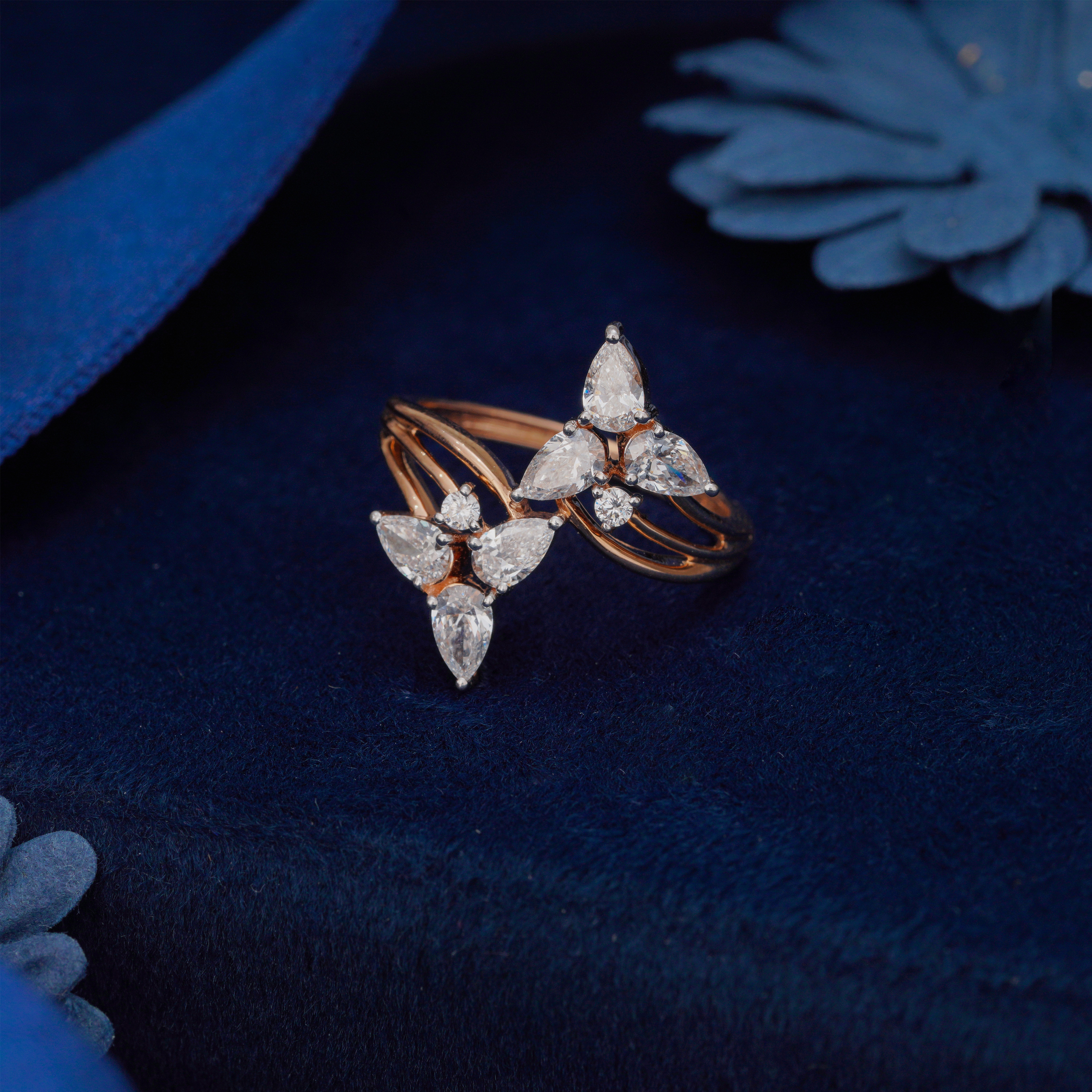 Rose gold ring with diamond accents on a dark blue fabric background.
Close-up of Aira floral wrap ring featuring pear-shaped lab grown diamonds in 18K yellow gold on a blue velvet background.
