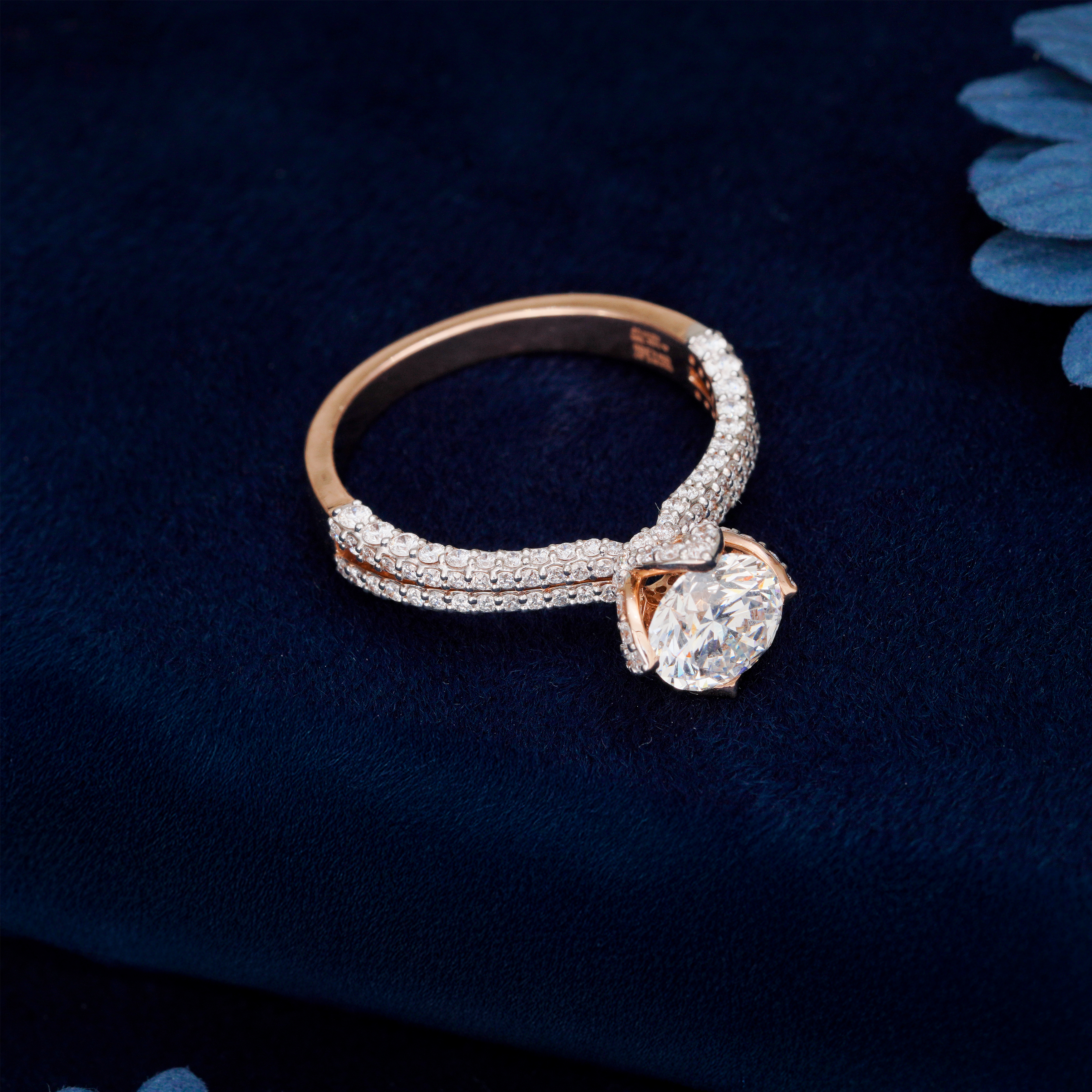 Rose gold ring with a diamond on a dark blue fabric background. Close-up of Aurelia round brilliant diamond engagement ring in yellow gold with split-shank pavé band