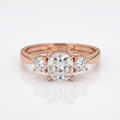 A rose gold three-stone engagement ring with a central cushion-cut diamond and two round side diamonds, displayed on a clean white reflective surface.