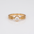 A yellow gold solitaire ring with a round-cut clear gemstone, featuring a wing-like twisted band design, shown on a clean white background with a soft reflection.