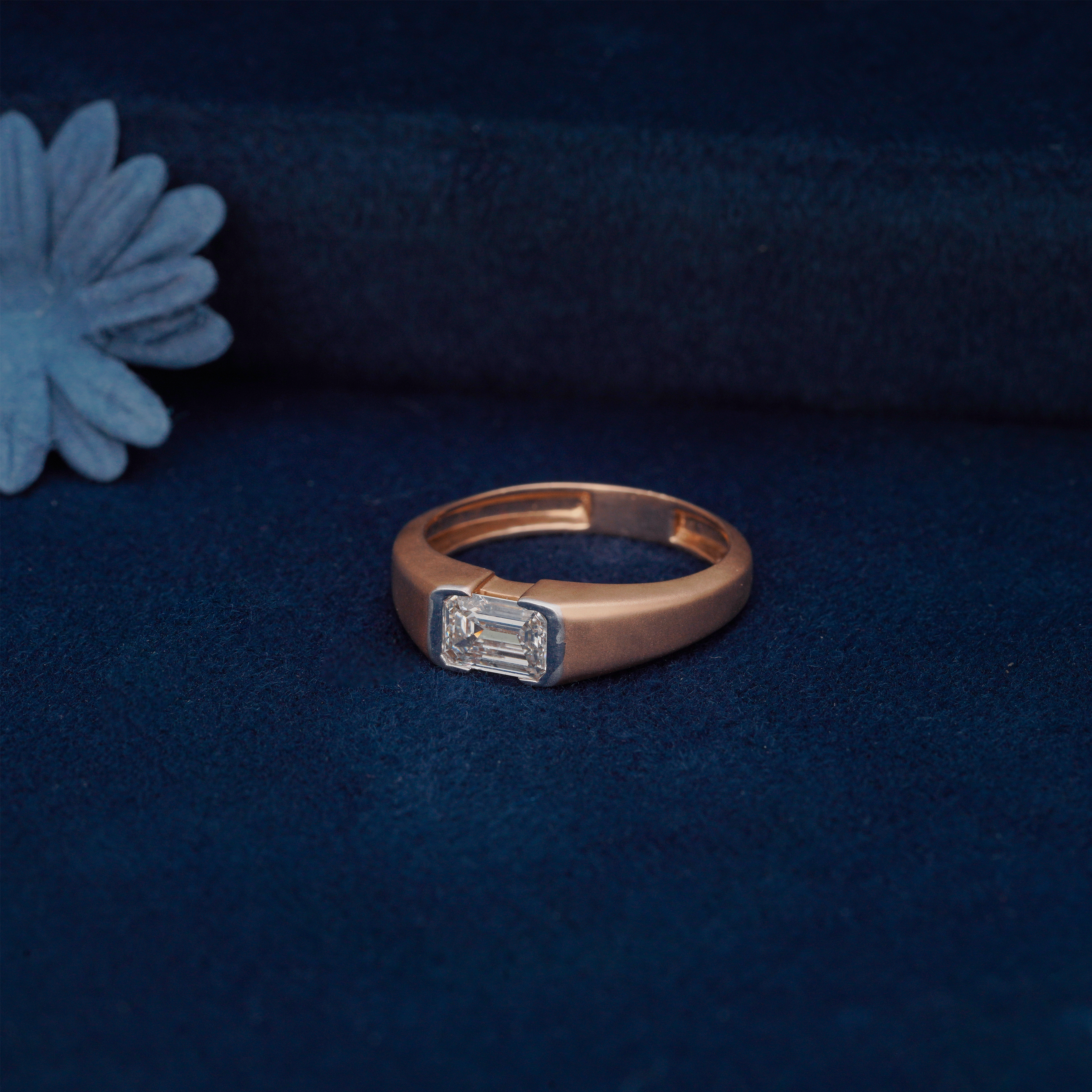 Single emerald cut lab diamond set in a wide, smooth yellow gold band, displayed on a deep blue velvet surface.