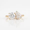 A pair of yellow gold engagement rings on a white reflective surface. One ring features a round brilliant-cut diamond, and the other features a pear-cut diamond, both set in gold prong settings.