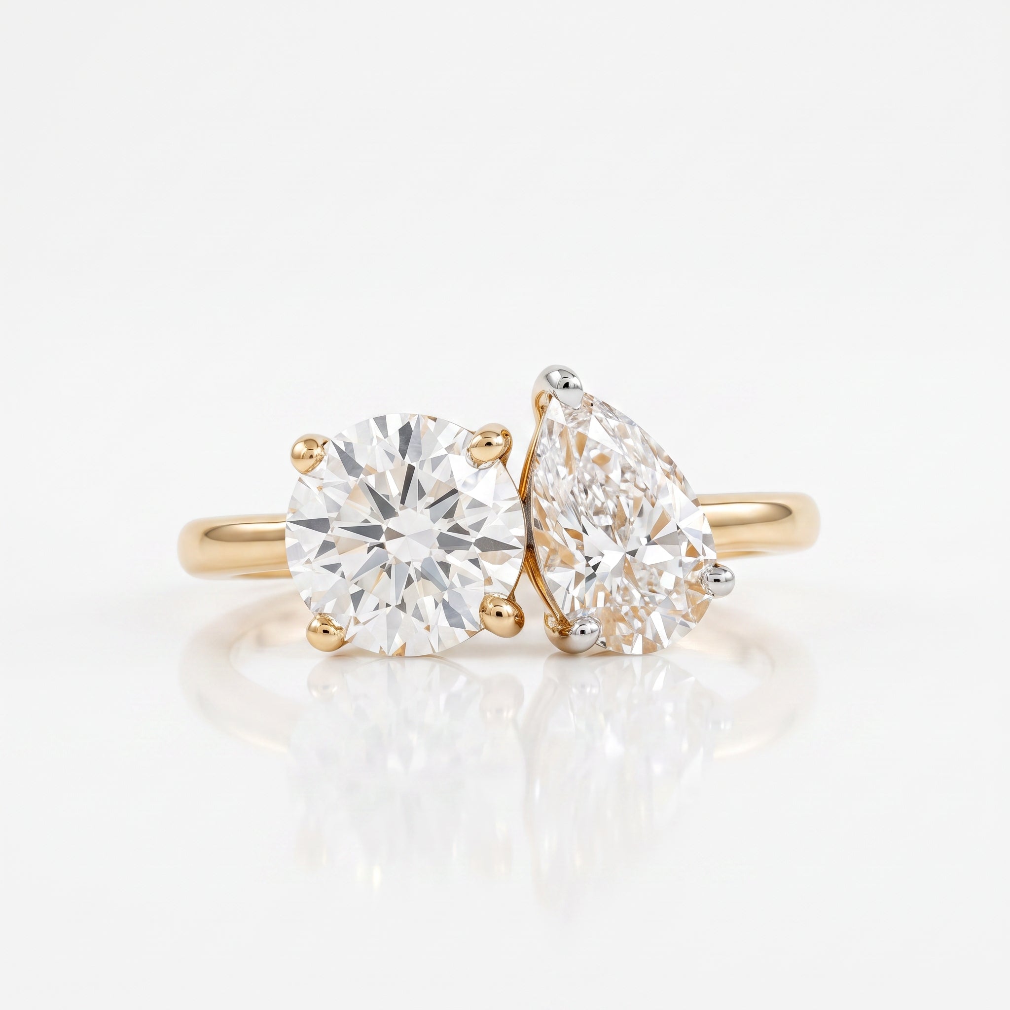 A pair of yellow gold engagement rings on a white reflective surface. One ring features a round brilliant-cut diamond, and the other features a pear-cut diamond, both set in gold prong settings.